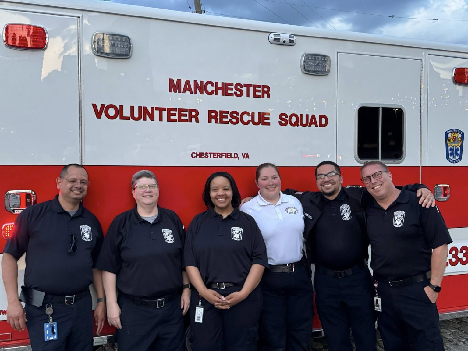 Six smiling members of the Manchester Volunteer Rescue Squad stand in front of their ambulance in Chesterfield, VA, wearing matching uniforms with identification badges.