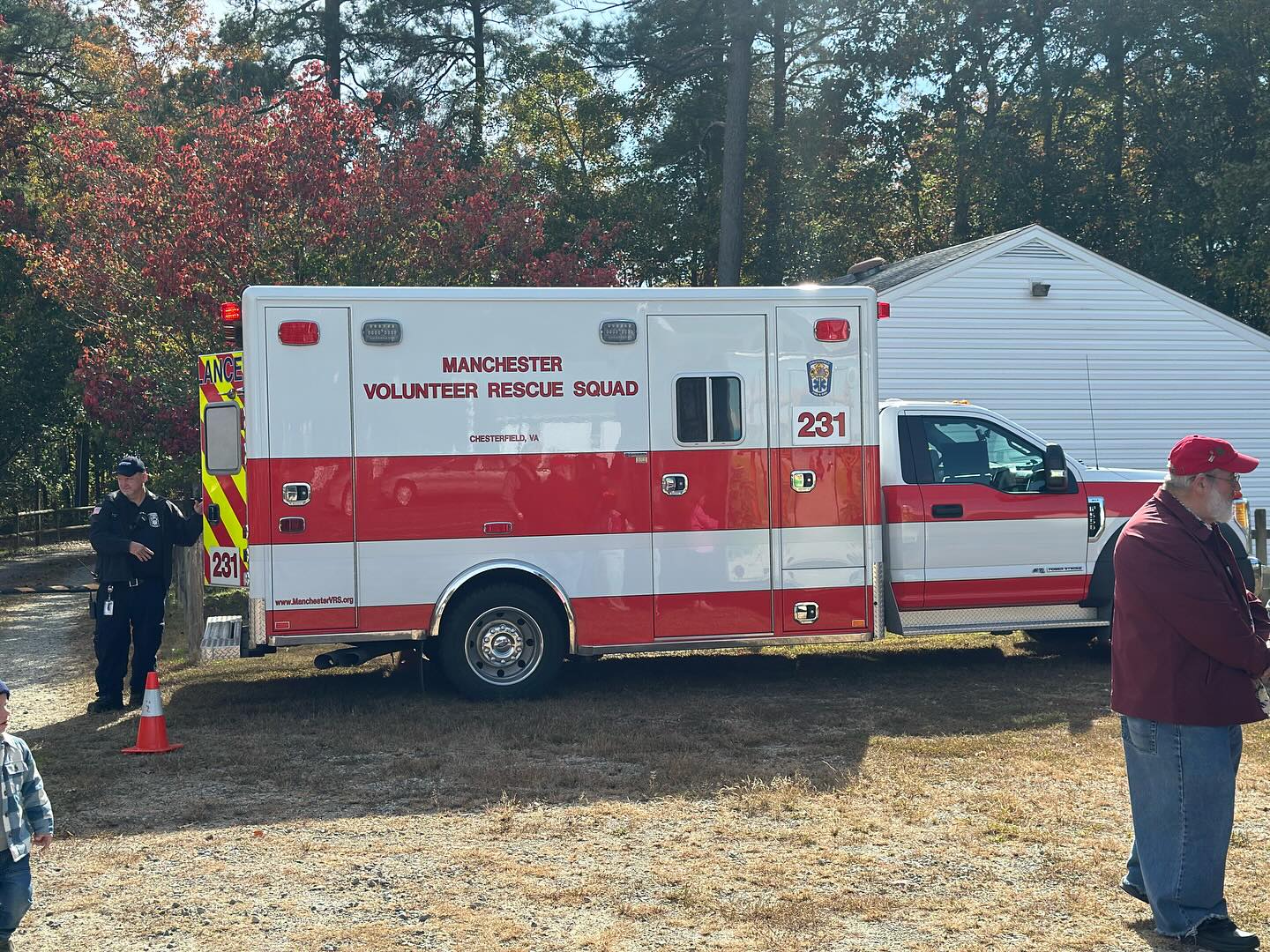 A red and white Manchester Volunteer Rescue Squad ambulance is parked on grass near a white building, with several people standing nearby under sunny skies and autumn trees.