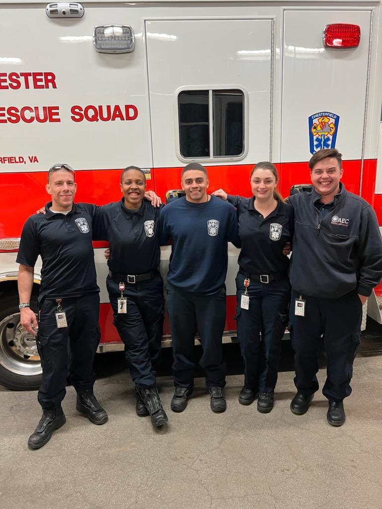 Five emergency medical personnel in uniform stand smiling in front of an ambulance with "Rescue Squad" text visible on the vehicle. They are standing close together with arms around each other.