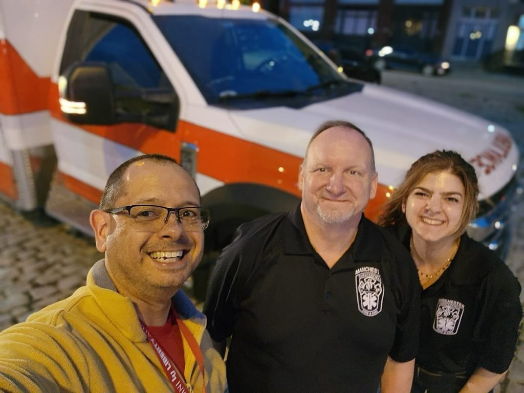 Three people smiling at the camera stand in front of an ambulance parked on a cobblestone street. Two wear black uniforms with medical emblems, and one wears glasses and a yellow jacket. It is evening or night.