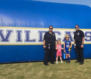 Two security officers stand with a young girl and a boy in sports gear in front of a large inflatable structure labeled "WILDCATS" on a sunny day.