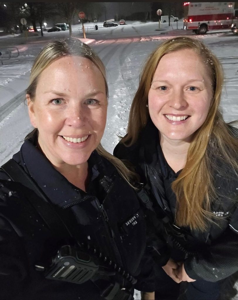 Two women in dark uniforms stand outside in the snow at night, smiling at the camera. A snow-covered street and a red emergency vehicle are visible in the background.