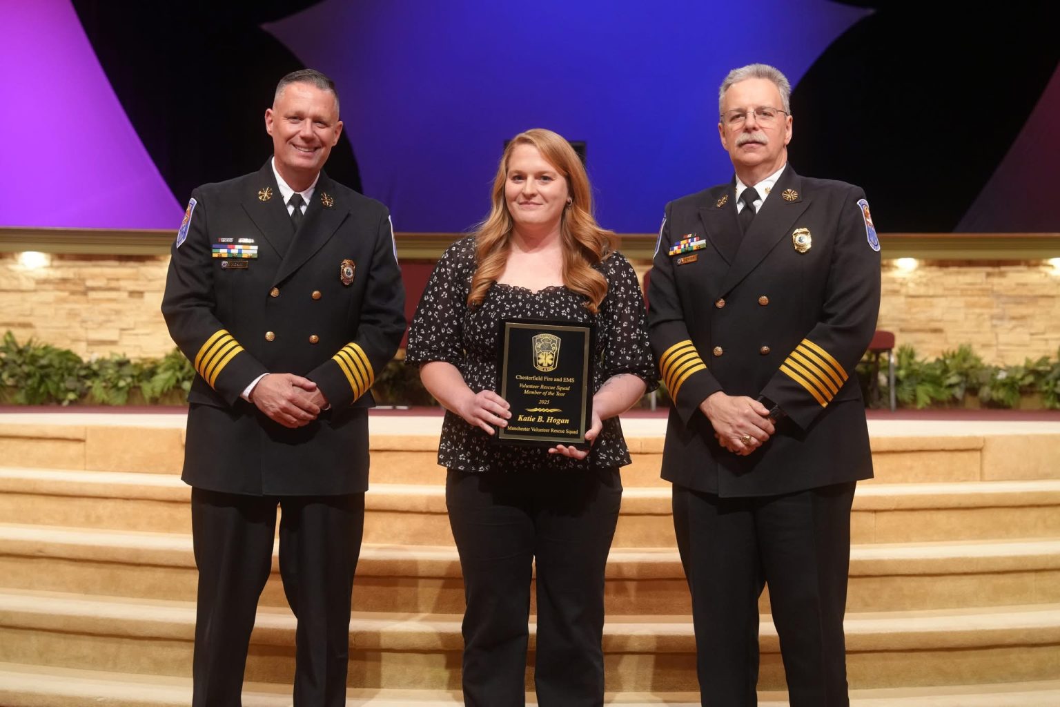 Three people pose on a stage; a woman in the center holds a plaque, flanked by two men in formal fire department uniforms, all facing the camera and smiling.